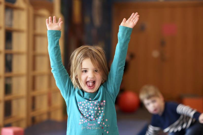 Smiling child in a teal shirt raising both arms in an indoor playroom.