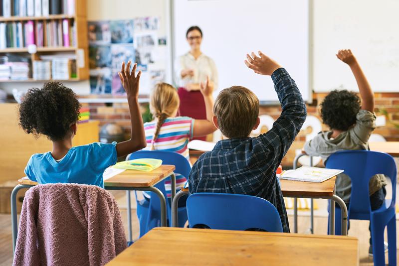 Seen from the back of a classroom, rows of young students raise their hands as their smiling teacher looks on.