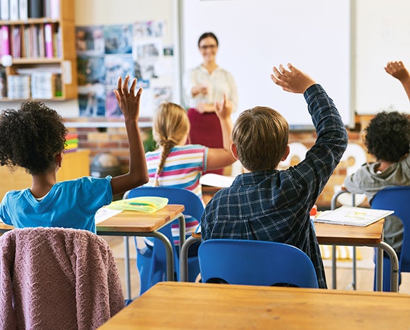 Seen from the back of a classroom, rows of young students raise their hands as their smiling teacher looks on.